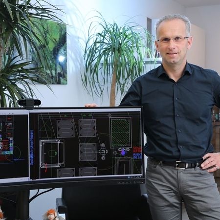 Michael Kolar posiert neben zwei Monitoren auf einem Schreibtisch